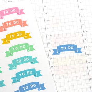 May include: Colorful "TO DO" banner stickers in pink, orange, yellow, green, teal, and blue. The stickers are on a white background with a grid pattern, and one blue sticker is placed on the grid. The stickers are ideal for planners and journals.