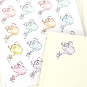 May include: A sticker sheet featuring multiple illustrations of watering cans in pastel shades of pink, blue, yellow, and orange, each depicted with water droplets. A cream-colored dot grid notebook is positioned in the foreground with a small bow logo.