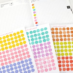 Könnte beinhalten: Ein offener Planer zeigt die Daten vom 27. und 28. März. Darunter befinden sich mehrere Blätter mit farbigen Punktaufklebern. Die Aufkleber sind in Gelb-, Rosa-, Blau-, Grün-, Orange- und Lilatönen gehalten. Der Markenname "SUGARLOOP" ist auf den Aufkleberbögen aufgedruckt.