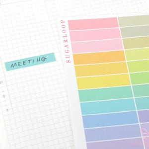 May include: A sheet of rectangular adhesive labels in a gradient of pastel colors including pink, orange, yellow, green, blue, and purple, with the text SUGARLOOP printed vertically in pink. A single blue label with the word MEETING is applied to a grid planner.
