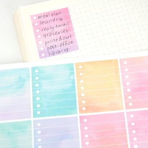 May include: A stack of white paper with a pink and purple sticky note that says "meal plan, laundry, reply email, groceries, print & cut, post-office, library." Below is a sheet of colorful watercolor-style planner stickers.