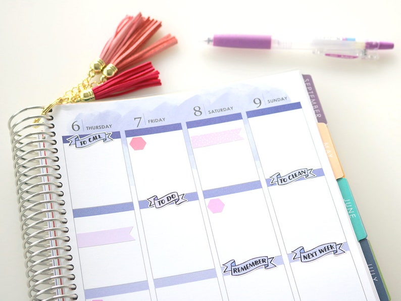 May include: A close-up of a planner with a silver coil binding. The planner has a light blue background with white and pink stickers. The stickers have text that reads "To Call", "To Do", "To Clean", "Remember", and "Next Week".