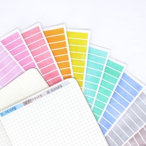 May include: A set of colorful washi tape with a watercolor effect. The tape is arranged in a fan shape with the colors ranging from pink to gray. The tape is labeled with the days of the week: Friday, Saturday, and Sunday.