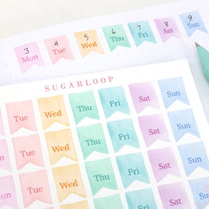 May include: A sheet of colorful watercolor planner stickers with the days of the week, labeled Mon through Sun. The stickers are in a variety of pastel colors, including pink, yellow, green, and blue. A teal pen rests on the page.