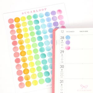 May include: A sheet of colourful watercolour dot stickers. The stickers are arranged in a rainbow pattern, with pink, orange, yellow, green, blue, and purple dots. The sheet is labelled "SUGARLOOP" in the top left corner.