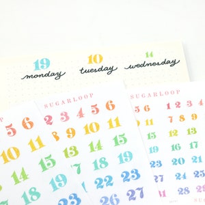 以下が含まれることがあります： 「Sugarloop」の文字と、月曜日、火曜日、水曜日の曜日が書かれた、カラフルな数字のステッカーのセット。数字はピンクから始まり青で終わる、虹色のグラデーションになっています。