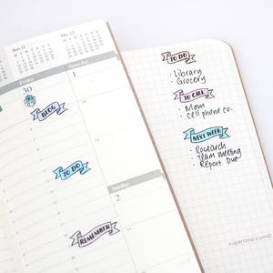 May include: A close-up of a planner with colorful stickers. The stickers are in the shape of banners and have text on them, such as "To Do", "Blog", and "To Call".