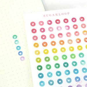 May include: A sheet of rainbow watercolor circle stickers with white heart cutouts. The stickers are arranged in rows and columns, with each row featuring a different color of the rainbow. The sheet is labeled "SUGARLOOP" at the top.