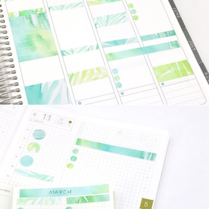 May include: A close-up of a planner with watercolor washi tape in green, blue, and yellow. The tape features a tropical leaf pattern. The planner has a gridded layout and is labeled "March".