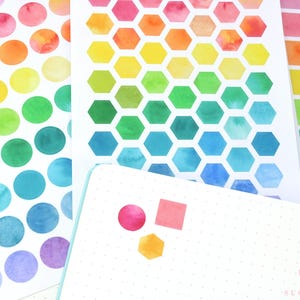 May include: A collection of colorful watercolor stickers in various shapes, including circles, hexagons, and squares. The stickers feature a rainbow of hues, from pink and orange to green and blue. A notebook with the text "sugarloop" is also visible.
