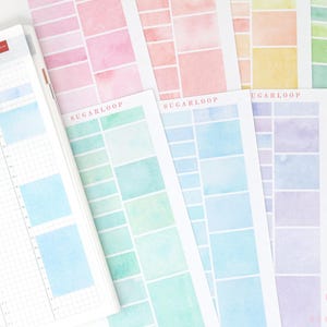 May include: A planner is open to a double-page spread, with a grid layout and blue watercolor blocks. Several sheets of watercolor paper with the word "SUGARLOOP" are visible, featuring colorful square and rectangular designs in pastel shades of pink, green, blue, and yellow.
