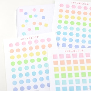 May include: A set of three sheets of pastel-coloured stickers with different shapes. The first sheet has circles in shades of pink, orange, yellow, green, blue, and purple. The second sheet has hexagons in the same colour scheme. The third sheet has squares in the same colour scheme. Each sheet has the word "Sugarloop" printed at the top.