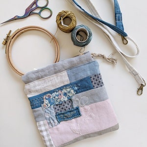 Può includere: Una borsa in tessuto patchwork con la scritta "MY PRECIOUS THINGS" è circondata da forniture per ricamo. Gli articoli includono un telaio da ricamo in legno, forbici, rocchetti di filo e un cordino in denim e lino. La borsa presenta tessuti blu, grigi e rosa.