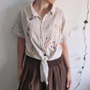 May include: A beige short-sleeved button-down shirt featuring delicate floral and botanical embroidery in various colours. The shirt has a collar, cuffed sleeves, and is tied in a knot at the front waist, paired with a dark brown skirt with an elastic waistband.