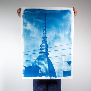 May include: A cyanotype print of a tall building with a spire, in shades of blue and white. The image is held up by a person. The building's details are visible against a cloudy sky. The print is on white paper.
