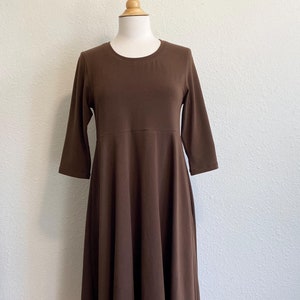 May include: A brown, knee-length dress with three-quarter sleeves is displayed on a mannequin. The dress has a fitted bodice and a flared skirt. The fabric appears to be a soft, solid color material. The dress is a solid brown color.