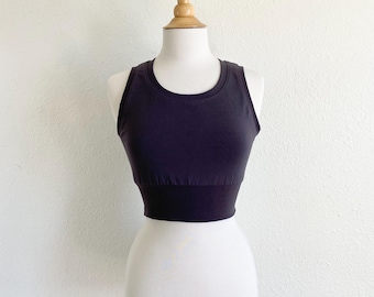 May include: A solid dark charcoal gray cropped tank top featuring a high rounded neckline and a wide, structured ribbed hem band that sits snugly at the waist. The garment is sleeveless with thick shoulder straps and a fitted silhouette.