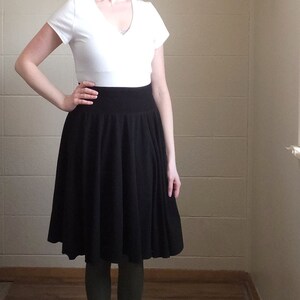 May include: A white short-sleeved V-neck top paired with a high-waisted black knee-length skirt featuring a wide waistband and a pleated, flared silhouette. The outfit is worn over dark olive green leggings, creating a layered appearance.