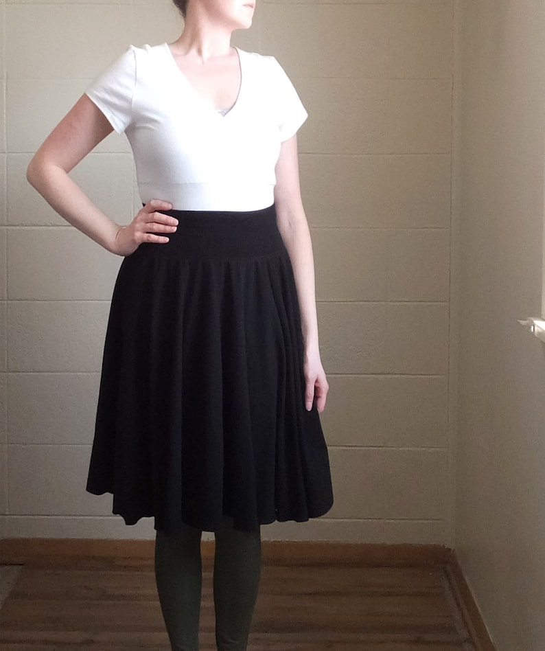 May include: A white short-sleeved V-neck top paired with a high-waisted black knee-length skirt featuring a wide waistband and a pleated, flared silhouette. The outfit is worn over dark olive green leggings, creating a layered appearance.