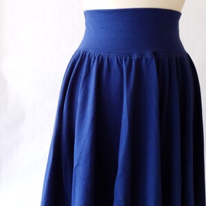 May include: A solid navy blue skirt featuring a wide, high-rise elasticized waistband and a gathered, flowing silhouette that falls from the waist. The garment is displayed on a mannequin, showcasing the deep blue fabric and pleated texture.