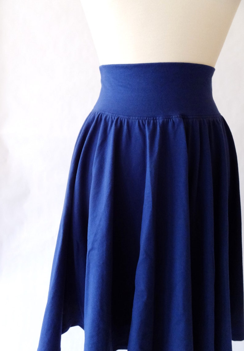 May include: A solid navy blue skirt featuring a wide, high-rise elasticized waistband and a gathered, flowing silhouette that falls from the waist. The garment is displayed on a mannequin, showcasing the deep blue fabric and pleated texture.