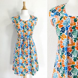 May include: A sleeveless dress with a colorful floral pattern. The dress features a scoop neckline, a tie waist, and a knee-length skirt. The floral design includes orange, blue, green, and white leaves. The dress is displayed on a mannequin.