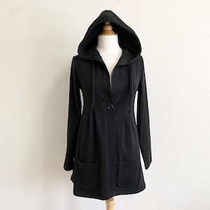 May include: A black hooded jacket with long sleeves and two front pockets. The jacket features a single button closure and a drawstring around the hood. The garment is displayed on a mannequin against a white background.