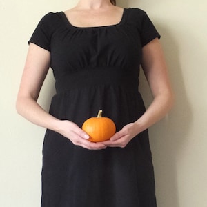 May include: A black dress with a square neckline and short sleeves. A small, round, orange pumpkin is held in front of the dress. The pumpkin has a green stem.