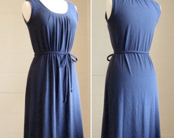 May include: A sleeveless navy blue maxi dress featuring a scoop neckline with subtle vertical pleating at the chest and a thin, matching fabric tie belt at the waist. The garment is displayed on a mannequin, showing both front and back views.