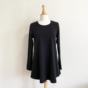 May include: A black long-sleeve tunic-style top with a rounded neckline and a flared hem. The garment is displayed on a wooden mannequin against a white background. The sleeves are long and fitted.