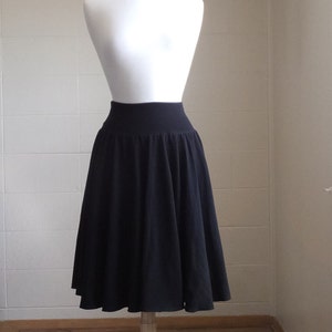 May include: A solid black knee-length skirt featuring a wide, fitted waistband and a flared, pleated silhouette. The skirt is displayed on a white mannequin, showcasing the smooth fabric texture and the consistent dark color throughout the garment.