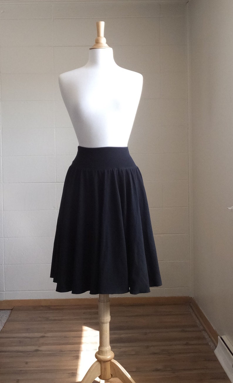 May include: A solid black knee-length skirt featuring a wide, fitted waistband and a flared, pleated silhouette. The skirt is displayed on a white mannequin, showcasing the smooth fabric texture and the consistent dark color throughout the garment.