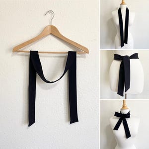 May include: A black tie-style scarf is displayed on a wooden hanger and styled on a mannequin in three different ways. The scarf is long and rectangular, with a smooth texture. The scarf can be worn as a necktie, belt, or bow.