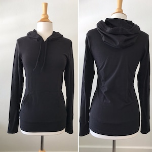 May include: A solid black long-sleeve hoodie featuring a front kangaroo pocket, a hood with matching black drawstrings, and fitted cuffs. The garment is displayed on a mannequin, showing both the front view with the pocket and the back view.