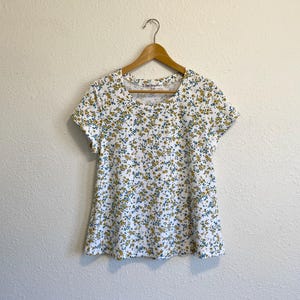 May include: White short-sleeved shirt with a floral print in shades of yellow and blue. The shirt has a round neckline and a relaxed fit.