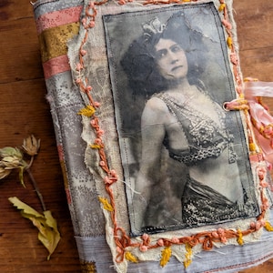 May include: A handmade journal with a vintage aesthetic. The cover features a black and white image of a woman in a decorative outfit. The journal is bound with fabric in shades of gray, pink, and yellow, and embellished with decorative stitching and a pink ribbon tie.