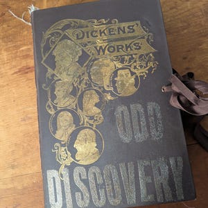 Puede incluir: Un libro marrón oscuro con letras doradas. El título es "Dickens' Works" y "Odd Discovery". La portada presenta un diseño de ilustraciones doradas de personas.