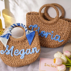 May include: Two round woven straw bags with light blue cursive lettering. One bag features the name Megan and a blue and white patterned scarf tied to the handle. The other bag features the name Gloria in blue yarn. Both have circular handles.