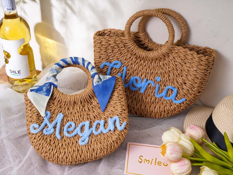 May include: Two round woven straw bags with light blue cursive lettering. One bag features the name Megan and a blue and white patterned scarf tied to the handle. The other bag features the name Gloria in blue yarn. Both have circular handles.