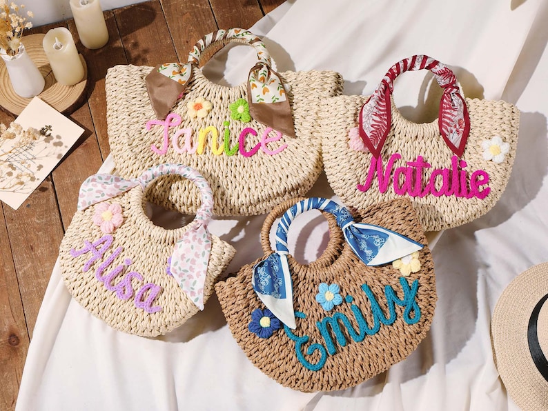 May include: Four woven straw handbags with rounded handles wrapped in patterned fabric scarves. Each bag features a personalized name in colorful yarn lettering and small fabric flower embellishments. The names displayed are Janice, Natalie, Alisa, and Emily.