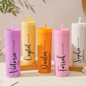 Puede incluir: Cinco vasos aislados de colores con pajitas. Los vasos son en tonos de morado, amarillo, naranja, rosa y blanco. Cada vaso tiene un nombre en fuente cursiva. El vaso morado dice "Victoria Bride". El vaso amarillo dice "Crystal". El vaso naranja dice "Danlen Maid of Honor". El vaso rosa dice "Farrah". El vaso blanco dice "Sophia Bridesmaid".
