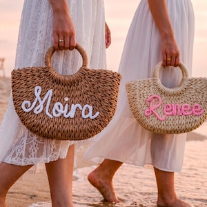May include: Two woven straw handbags with round handles. The darker brown bag features the name Moira in white script lettering, while the lighter beige bag displays the name Renee in pink script lettering, both crafted from textured material.