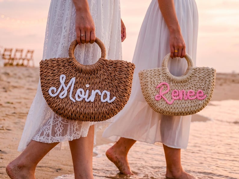 May include: Two woven straw handbags with round handles. The darker brown bag features the name Moira in white script lettering, while the lighter beige bag displays the name Renee in pink script lettering, both crafted from textured material.
