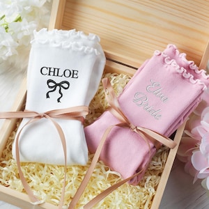 Może przedstawiać: Drewniane pudełko zawiera dwie pary skarpet. Jedna para jest biała z napisem "CHLOE" i czarną kokardą. Druga para jest różowa z napisem "Elva Bride" wyszytym srebrną nicią. Obie pary są związane jasnobrązową wstążką.