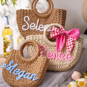 May include: Three woven straw tote bags with round handles. The back bag is brown with the white embroidered name "Selena". The middle bag is light beige with the pink embroidered name "Renee" and a pink gingham bow. The front bag is brown with the light blue embroidered name "Megan".