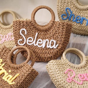 May include: A collection of five woven straw handbags with round handles, each featuring a personalized name embroidered in cursive yarn. The names displayed on the bags are Crystal in pink, Selena in white, Sherry in blue, Alrial in yellow, and Renee in pink.
