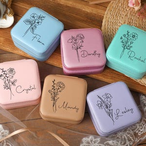 Peut inclure: Six bo&icirc;tes &agrave; bijoux carr&eacute;es de couleurs pastel : bleu clair, rose, vert menthe, p&ecirc;che, beige et lavande. Chaque bo&icirc;te pr&eacute;sente un motif floral et un nom en &eacute;criture cursive. Les bo&icirc;tes sont dispos&eacute;es sur une surface en bois.