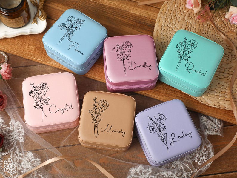 Peut inclure: Six bo&icirc;tes &agrave; bijoux carr&eacute;es de couleurs pastel : bleu clair, rose, vert menthe, p&ecirc;che, beige et lavande. Chaque bo&icirc;te pr&eacute;sente un motif floral et un nom en &eacute;criture cursive. Les bo&icirc;tes sont dispos&eacute;es sur une surface en bois.