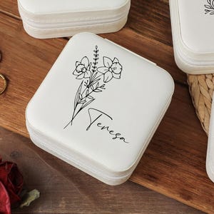 Peut inclure: Bo&icirc;te &agrave; bijoux carr&eacute;e blanche avec fermeture &eacute;clair, orn&eacute;e d'un motif floral noir et du nom "Teresa". Deux bagues sont visibles sur une surface en bois. D'autres bo&icirc;tes similaires sont en arri&egrave;re-plan.