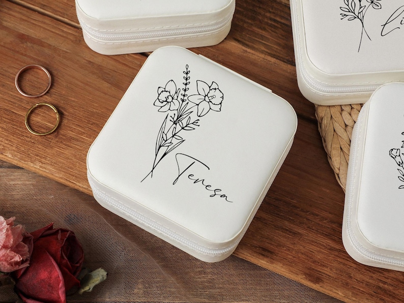 Peut inclure: Bo&icirc;te &agrave; bijoux carr&eacute;e blanche avec fermeture &eacute;clair, orn&eacute;e d'un motif floral noir et du nom "Teresa". Deux bagues sont visibles sur une surface en bois. D'autres bo&icirc;tes similaires sont en arri&egrave;re-plan.